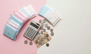 Foto ilustrasi pembalut wanita bersanding dengan koin dan kalkulator, menyimbolkan beban ekonomi dari pengenaan pajak (Pink Tax) pada kebutuhan dasar perempuan.