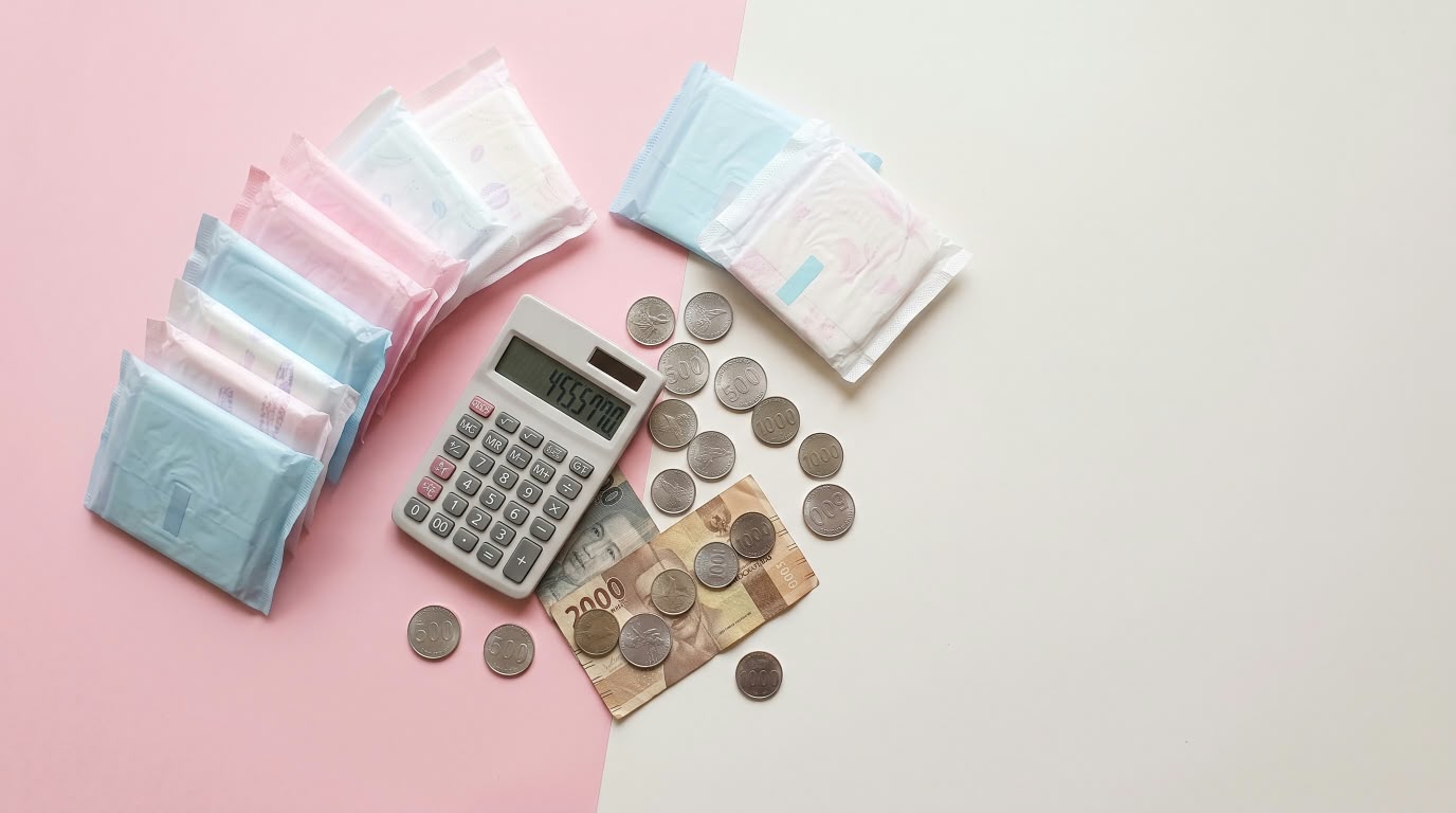 Foto ilustrasi pembalut wanita bersanding dengan koin dan kalkulator, menyimbolkan beban ekonomi dari pengenaan pajak (Pink Tax) pada kebutuhan dasar perempuan.